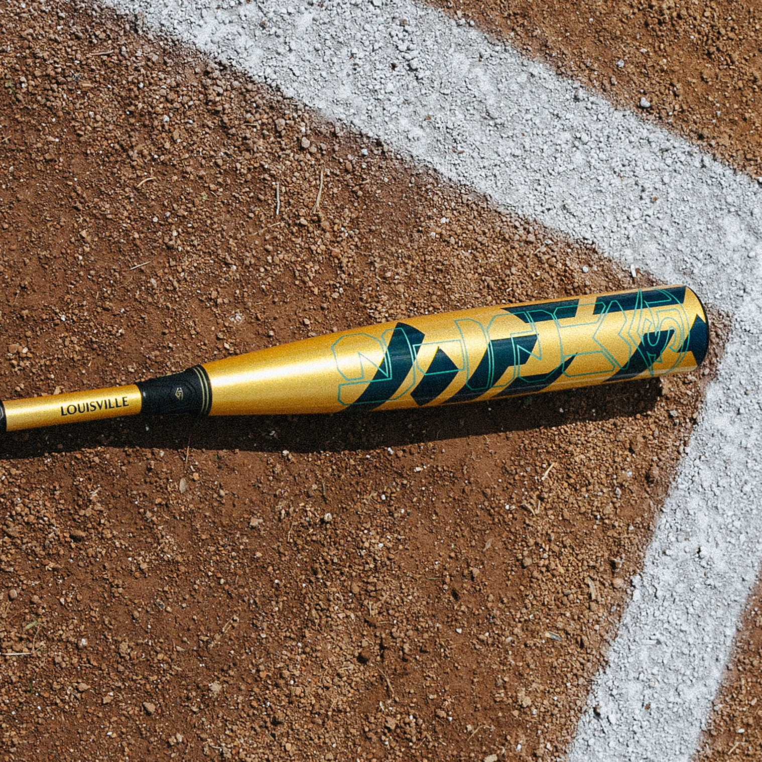 A gold and green Louisville Slugger baseball bat lies on a dirt field near a white chalk baseline.