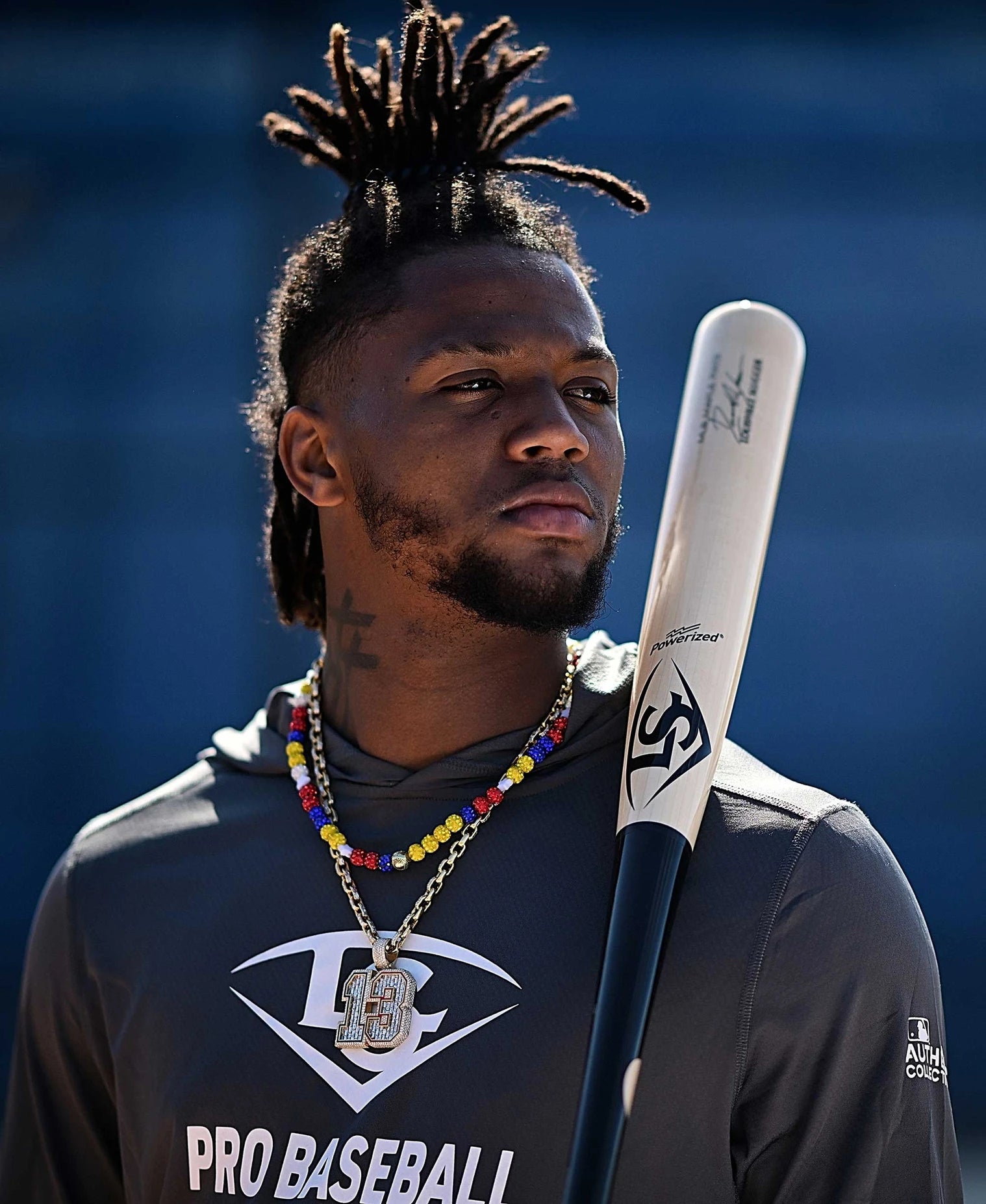 A baseball player wearing a gray Pro Baseball hoodie and colorful beaded necklaces holds a Louisville Slugger Pro Prime Signature Series RA13 Ronald Acuna Jr. Game Model Wood Baseball Bat (WBL2934010) over his shoulder, looking off into the distance with a serious expression.