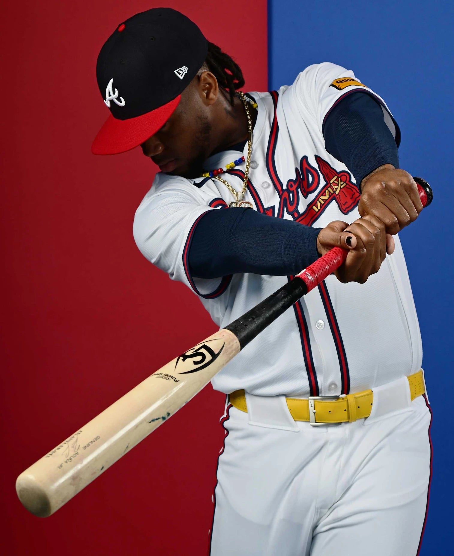 A baseball player in a white Atlanta Braves uniform and red cap holds a Louisville Slugger Pro Prime Signature Series RA13 Ronald Acuna Jr. Game Model Wood Baseball Bat (WBL2934010), posed as if swinging. The background is split blue and red, with the player’s face mostly hidden beneath the cap.