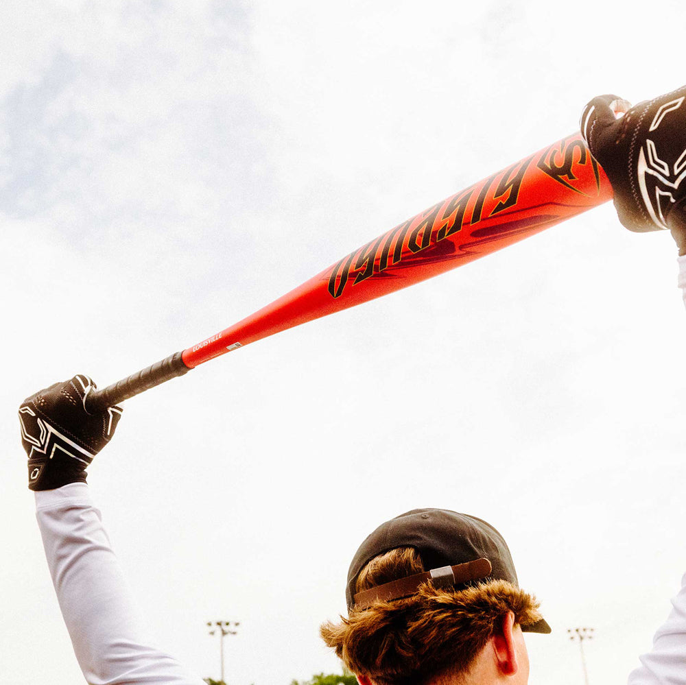 A person in a black cap and gloves holds the 2025 Louisville Slugger Dynasty (-3) BBCOR Baseball Bat (WBL2969010) high above their head, set against a cloudy sky with blurred outdoor lights in the background.