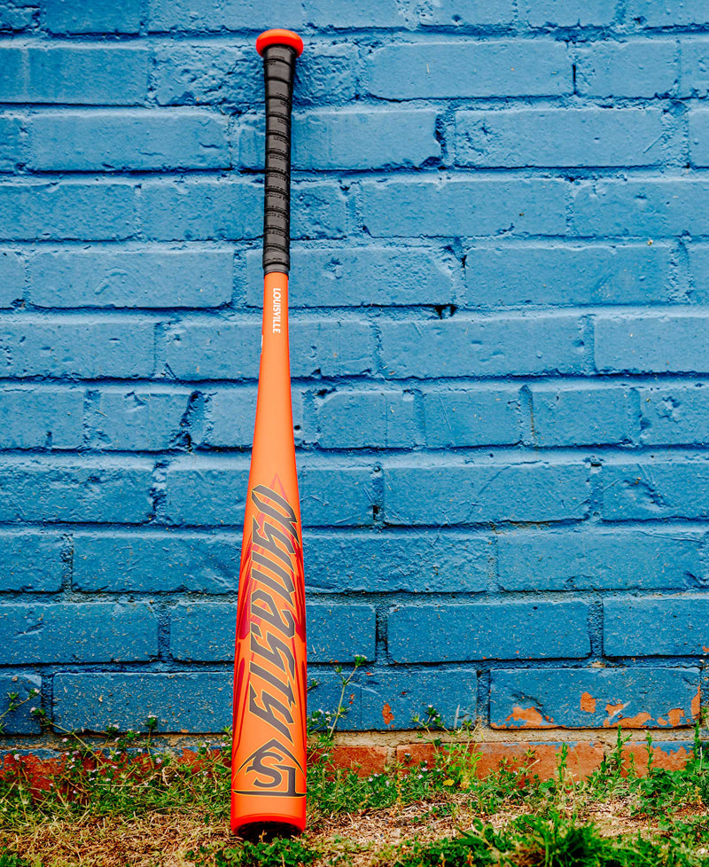 The 2025 Louisville Slugger Dynasty (-3) BBCOR Baseball Bat (WBL2969010), in orange with a black grip and lettering, stands upright against a blue brick wall, resting on grass and dirt.