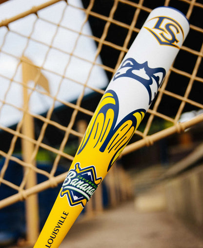 A close-up of the 2025 Louisville Slugger Savannah Bananas JBB (-10) USSSA Baseball Bat (WBL4006010), featuring a banana graphic, Savannah Bananas text, and Louisville Slugger branding against a tan net with a blurred stadium backdrop.