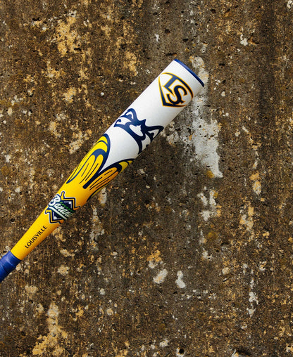 A 2025 Louisville Slugger Savannah Bananas JBB (-10) USSSA Baseball Bat, featuring yellow, blue, and white colors with team logos, rests against a weathered brown concrete wall.