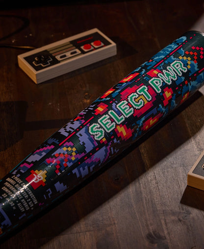 A vibrant 2026 Louisville Slugger Select PWR Player 1 (-3) BBCOR Baseball Bat (WBL4182010) with a pixelated design sits on a dark wooden table beside a retro Nintendo controller.