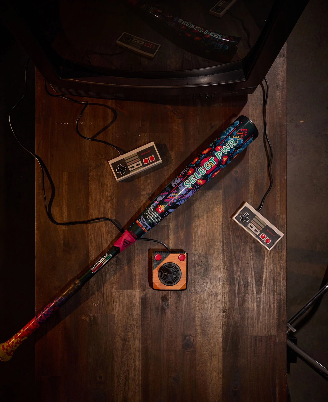 A 2026 Louisville Slugger Select PWR Player 1 (-3) BBCOR Baseball Bat rests beside two NES controllers and a vintage camera on a wooden table, all positioned next to a classic CRT television.
