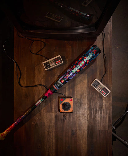 A 2026 Louisville Slugger Select PWR Player 1 (-3) BBCOR Baseball Bat rests beside two NES controllers and a vintage camera on a wooden table, all positioned next to a classic CRT television.