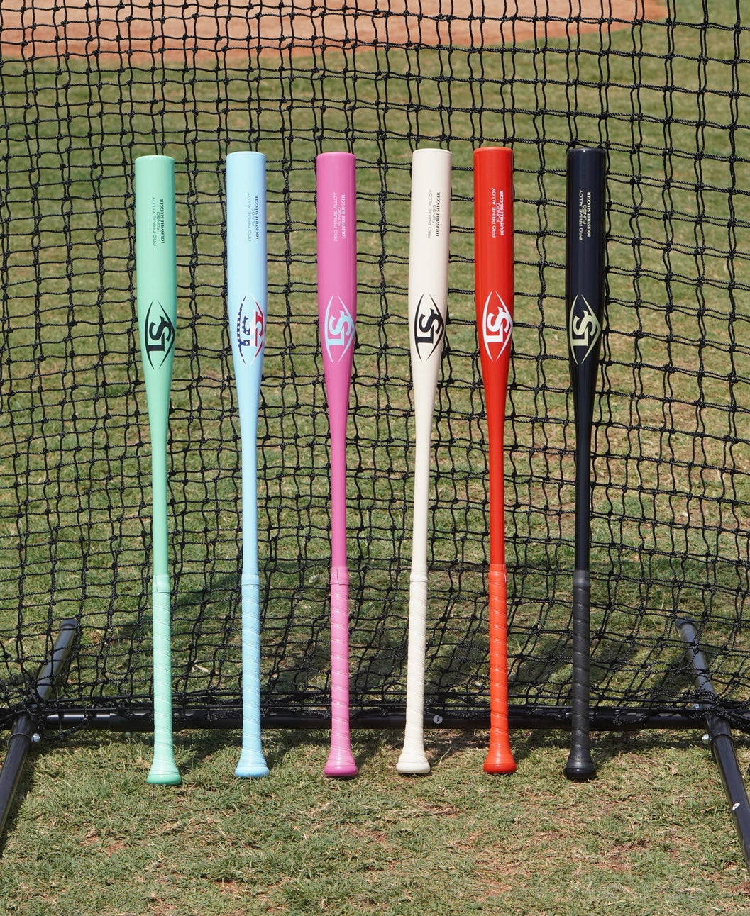 Six colorful baseball bats—mint green, sky blue, pink, cream, red, and black—stand upright in front of a net on a grassy field, with the infield dirt visible in the background. Among them is the Louisville Slugger Pro Prime Alloy Fungo (-13) 35″ Black Training Bat (WBL42010102235), a specialized training bat from Louisville Slugger designed for coaches to perfect their skills during practice.