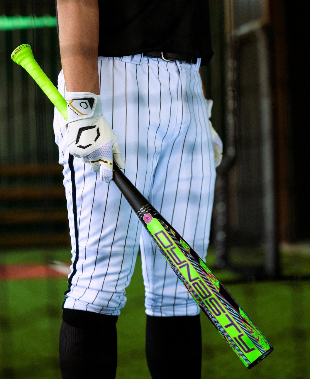 A baseball player in white pinstripe pants and gloves holds a neon green and black 2027 Louisville Slugger Dynasty (-3) BBCOR Baseball Bat (WBL4250010) while standing on turf.