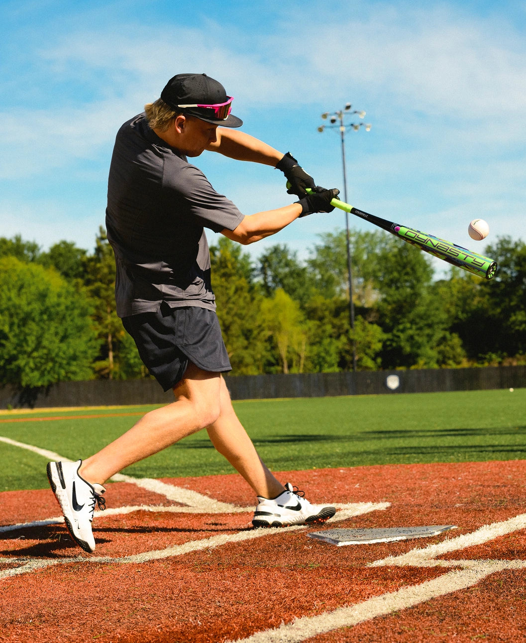Dressed in sportswear, a person swings a Louisville Slugger 2027 Dynasty (-3) BBCOR Baseball Bat (WBL4250010) at a pitched baseball on an outdoor field with vibrant green turf and trees in the background.