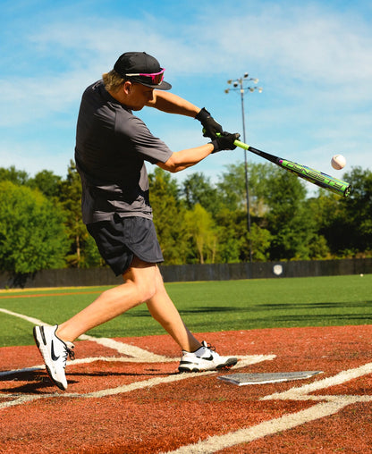 Dressed in sportswear, a person swings a Louisville Slugger 2027 Dynasty (-3) BBCOR Baseball Bat (WBL4250010) at a pitched baseball on an outdoor field with vibrant green turf and trees in the background.