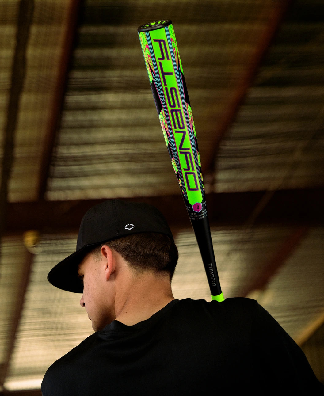 A person in a black shirt and cap holds the vibrant 2027 Louisville Slugger Dynasty (-3) BBCOR Baseball Bat (WBL4250010) over their shoulder in an indoor setting with a metal roof.