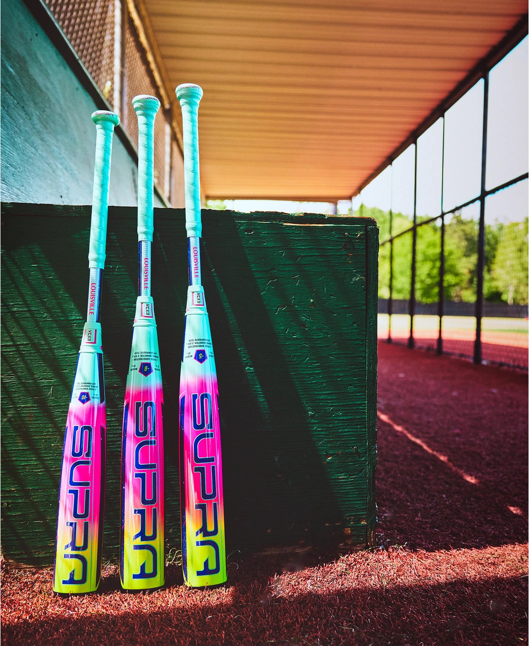 The Louisville Slugger 2027 Supra (-10) USSSA bat (WBL4251010) stands upright by a green dugout wall on red dirt, showing off its teal handle, vibrant pink, purple, and yellow design, and advanced APX Composite Technology.