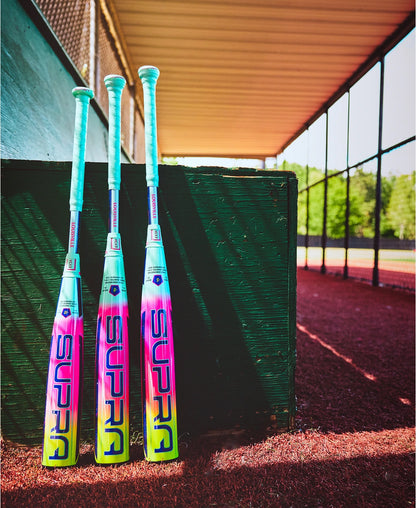The Louisville Slugger 2027 Supra (-10) USSSA bat (WBL4251010) stands upright by a green dugout wall on red dirt, showing off its teal handle, vibrant pink, purple, and yellow design, and advanced APX Composite Technology.
