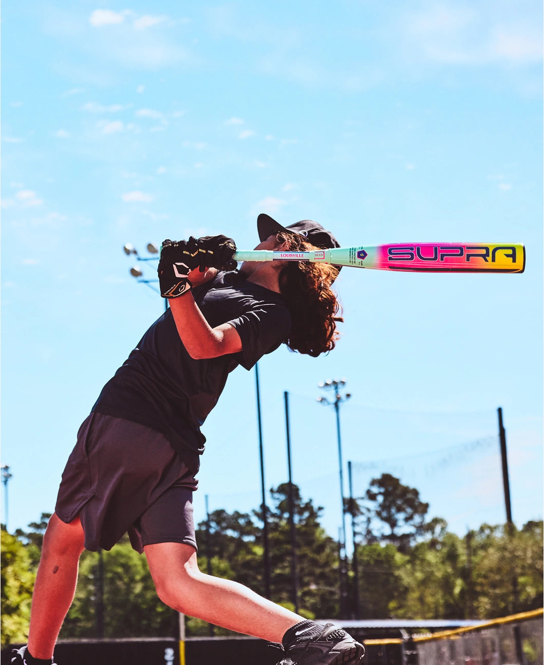 A young ballplayer in a dark uniform swings a 2027 Louisville Slugger Supra (-8) 2 3/4" USSSA Baseball Bat (WBL4252010) with composite tech on a sunny outdoor field, amid trees and light poles.