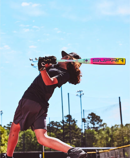 A young ballplayer in a dark uniform swings a 2027 Louisville Slugger Supra (-8) 2 3/4" USSSA Baseball Bat (WBL4252010) with composite tech on a sunny outdoor field, amid trees and light poles.