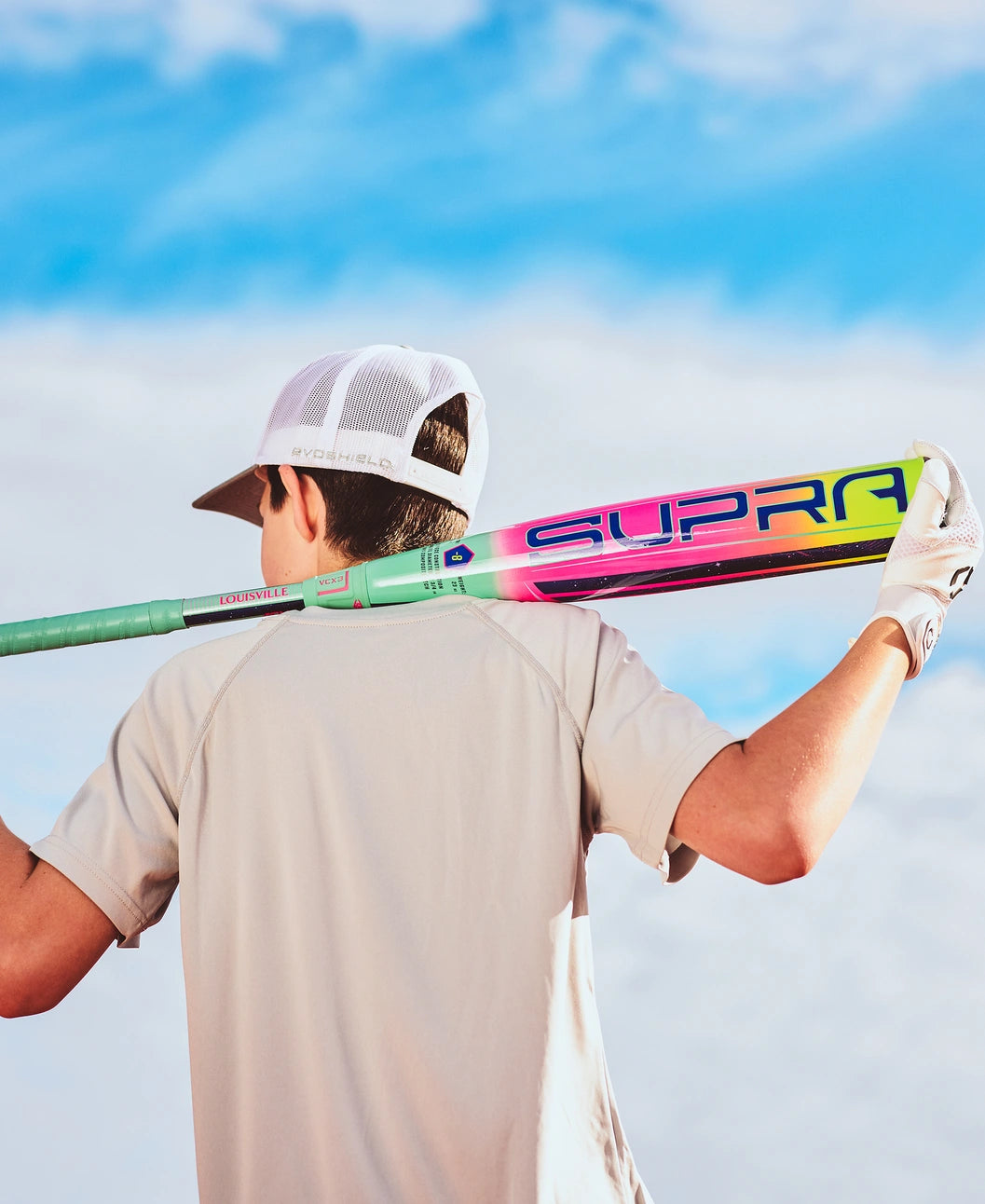 A person in a white cap and glove holds a colorful Louisville Slugger 2027 Supra (-8) USSSA Baseball Bat (WBL4252010) over their shoulders, facing away from the camera against a bright blue sky.