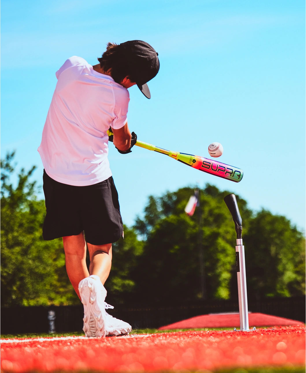 A child swings the 2027 Louisville Slugger Supra (-10) 2 5/8" USA Baseball Bat (WBL4258010) at a ball on a tee outdoors, with green trees and a blue sky in the background.
