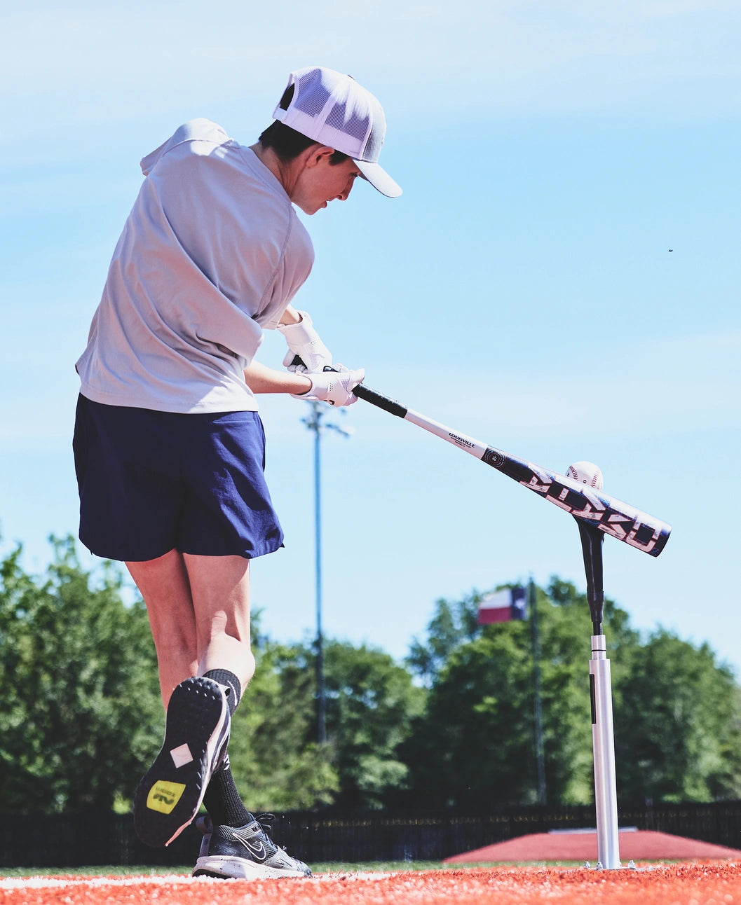 A person wearing a white hat and shirt swings the Louisville Slugger 2027 Omaha (-10) 2 5/8″ USA Baseball Bat (WBL4262010) at a ball on a tee on a sunny day, with trees and a flag in the background.