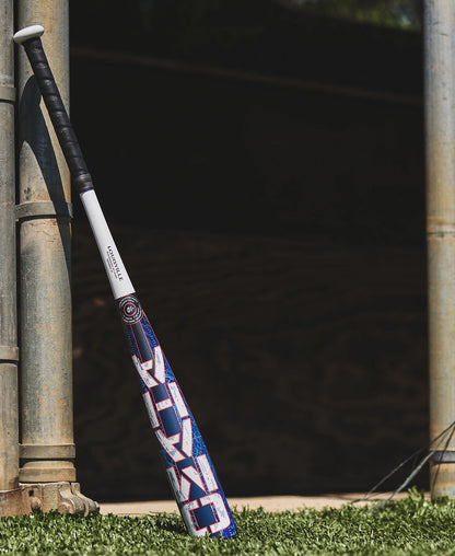 A Louisville Slugger 2027 Omaha (-10) 2 5/8″ USA Baseball Bat (WBL4262010), featuring a balanced swing weight and one-piece alloy construction with a blue, red, and white design, leans against a metal pole on green turf beside a shaded dugout area.