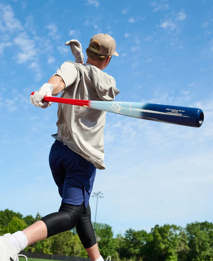 On a sunny day, a baseball player takes the field with his Louisville Slugger Pro Prime RA13 USA Maple Wood Baseball Bat (WBL4296010), featuring the Professional Series Finish. Wearing a cap, t-shirt, shorts, leggings, and a glove—his gear enhanced by Seamless Decals for a polished appearance—he plays beneath trees and blue sky.