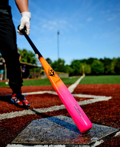 A person in baseball gear stands at home plate holding a vibrant pink and orange Louisville Slugger Pro Prime RA13 Sunset Maple Wood Baseball Bat (WBL4297010) on a sunny day, with a blurred green background and blue sky visible, highlighting its Professional Series Finish.