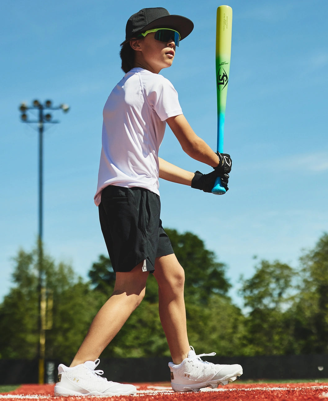On a sunny baseball field with trees and a light pole in the background, a young boy dressed in sportswear stands ready to bat, channeling Ronald Acuña Jr.’s style with his sunglasses, cap, and gloves. He grips the Louisville Slugger Youth Prime RA13 Ronald Acuña Jr. Maple Wood Baseball Bat (WBL4298010), its eye-catching neon tricolor fade reflecting his energy and passion for the game.