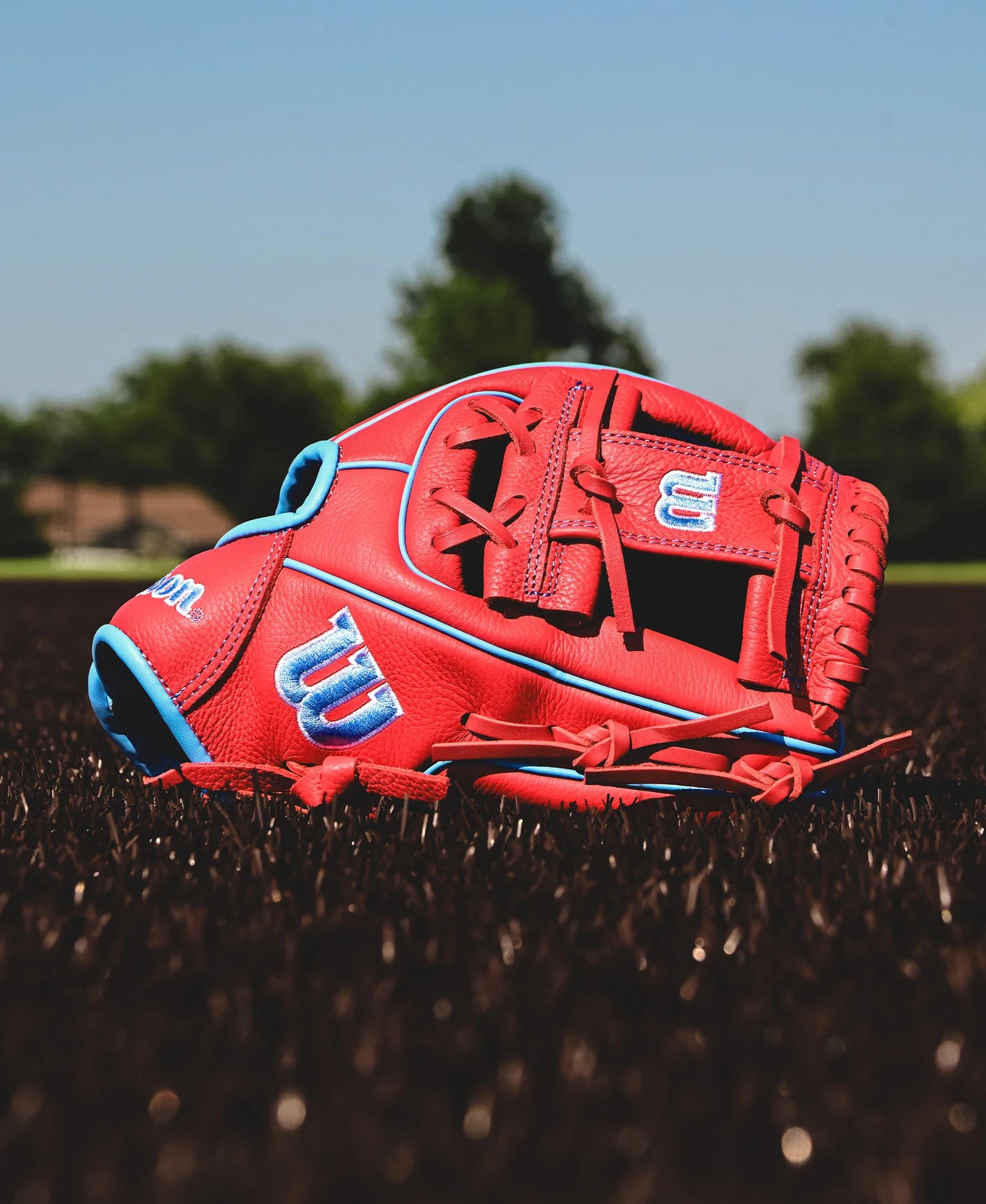 A Wilson A500 11.5" Baseball Glove: WBW102540115 by Wilson, featuring blue accents, lies on brown turf under a clear blue sky with green trees in the background.
