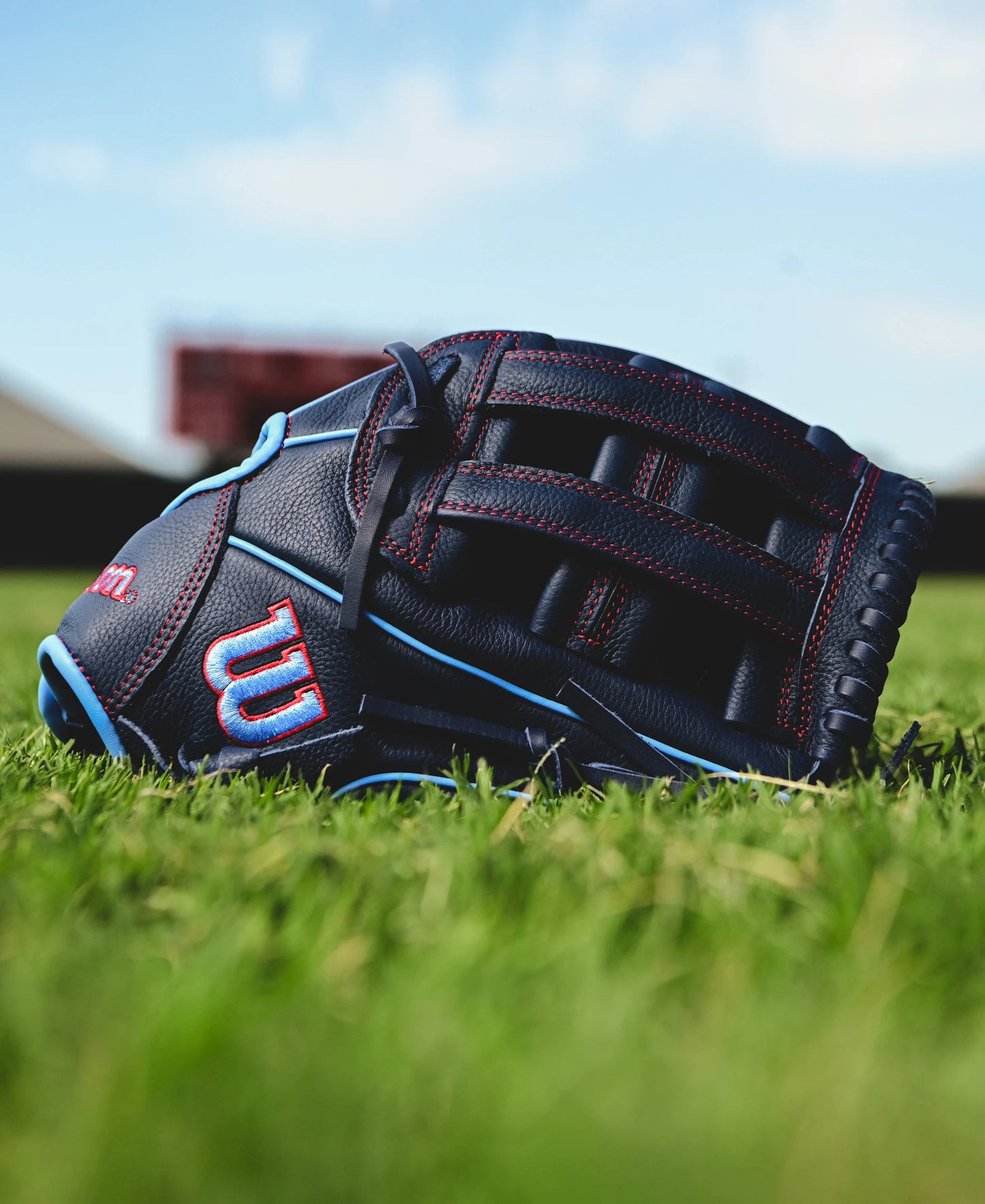 A close-up of the Wilson A500 12" Baseball Glove (WBW10254212) in black and blue with red stitching, resting on green grass, with a blurred scoreboard and sky in the background.