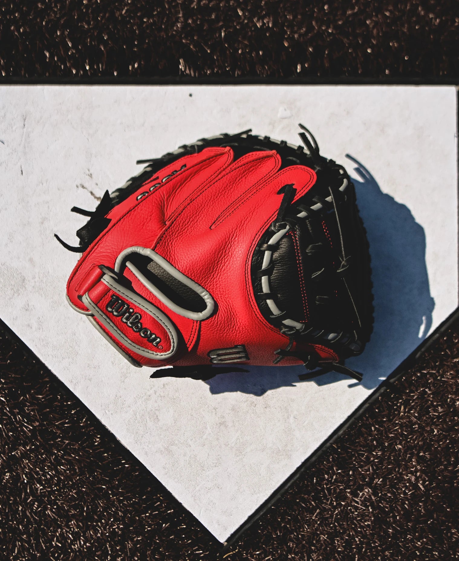 A Wilson A500 32" Baseball Catcher's Mitt (WBW10254632) by Wilson rests on home plate, its red color casting a shadow across the white surface and standing out against the dark brown dirt around the plate.