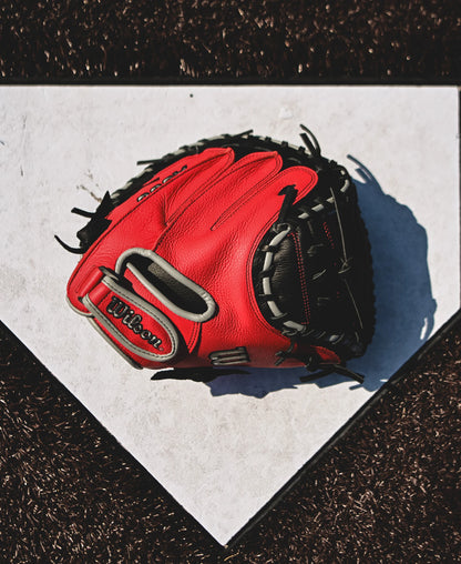 A Wilson A500 32" Baseball Catcher's Mitt (WBW10254632) by Wilson rests on home plate, its red color casting a shadow across the white surface and standing out against the dark brown dirt around the plate.
