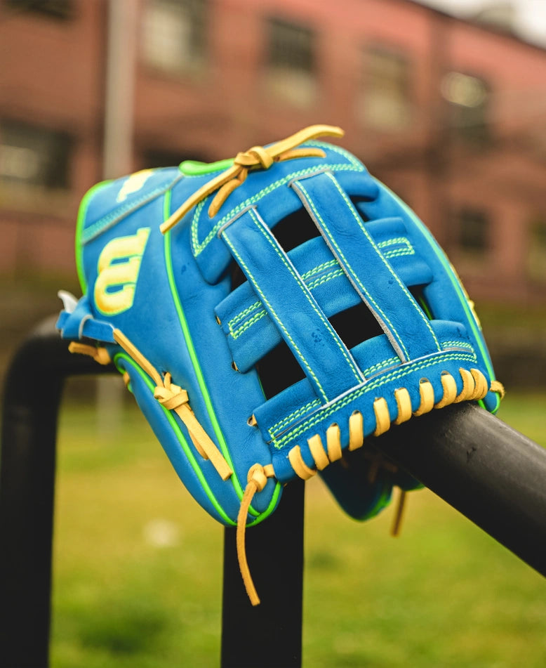 A Wilson A2000 PP05 11.5" Baseball Glove (SPRING 2025: WBW102548115) in blue and yellow, crafted with Pro Stock® leather, sits on a black rail outdoors, with green grass and a blurred building in the background.