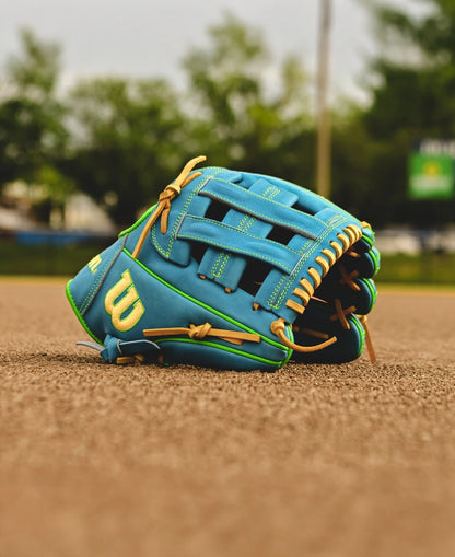 A Wilson A2000 PP05 11.5" Baseball Glove (SPRING 2025: WBW102548115) in blue and tan with yellow stitching rests on a baseball field’s dirt, while trees and a scoreboard blur softly in the background.
