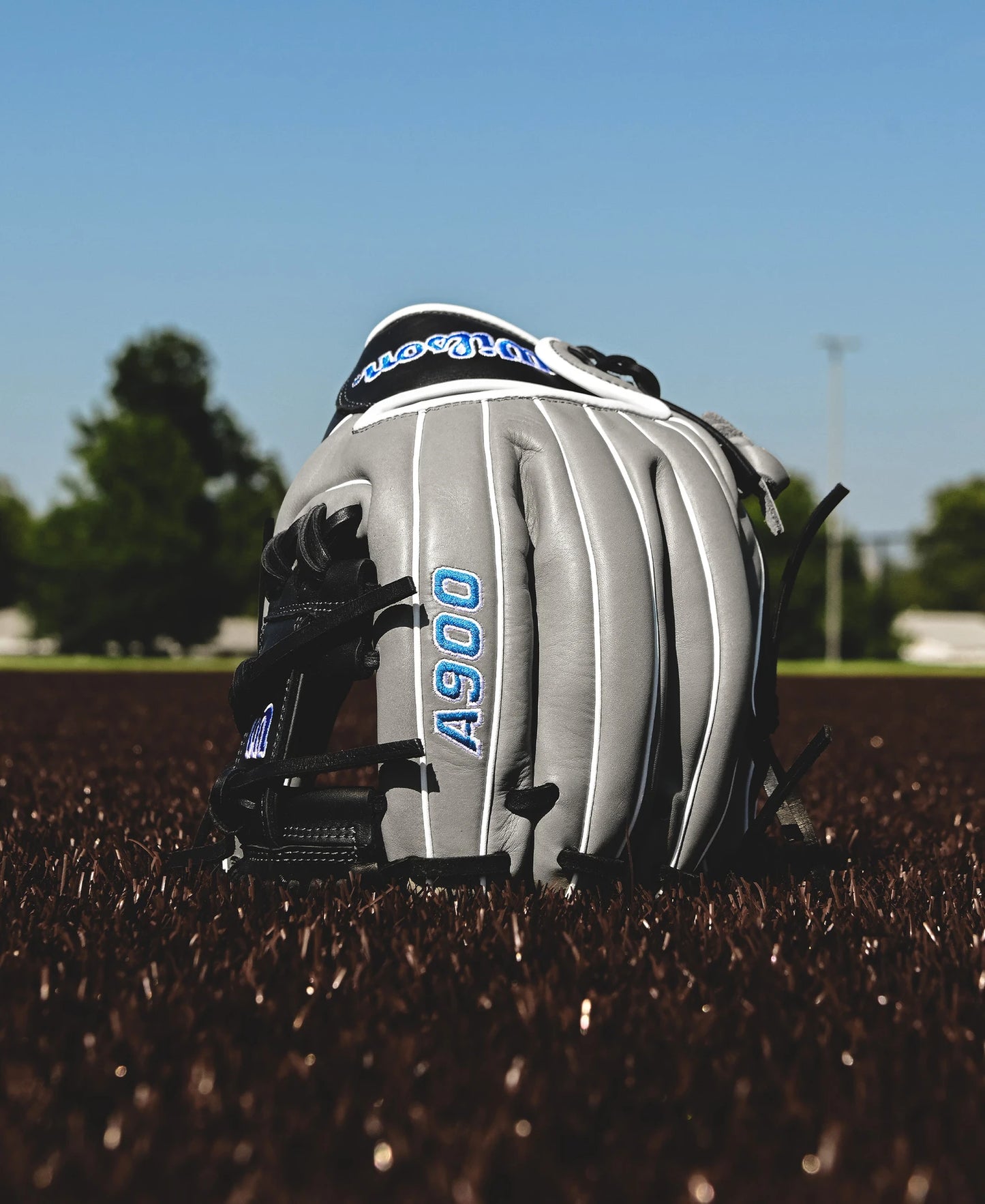 A Wilson A900 PF1125 11.25" youth baseball glove by Wilson, in gray and black with "A900" stitched in blue, rests on a brown field with green trees and blue sky blurred in the background.