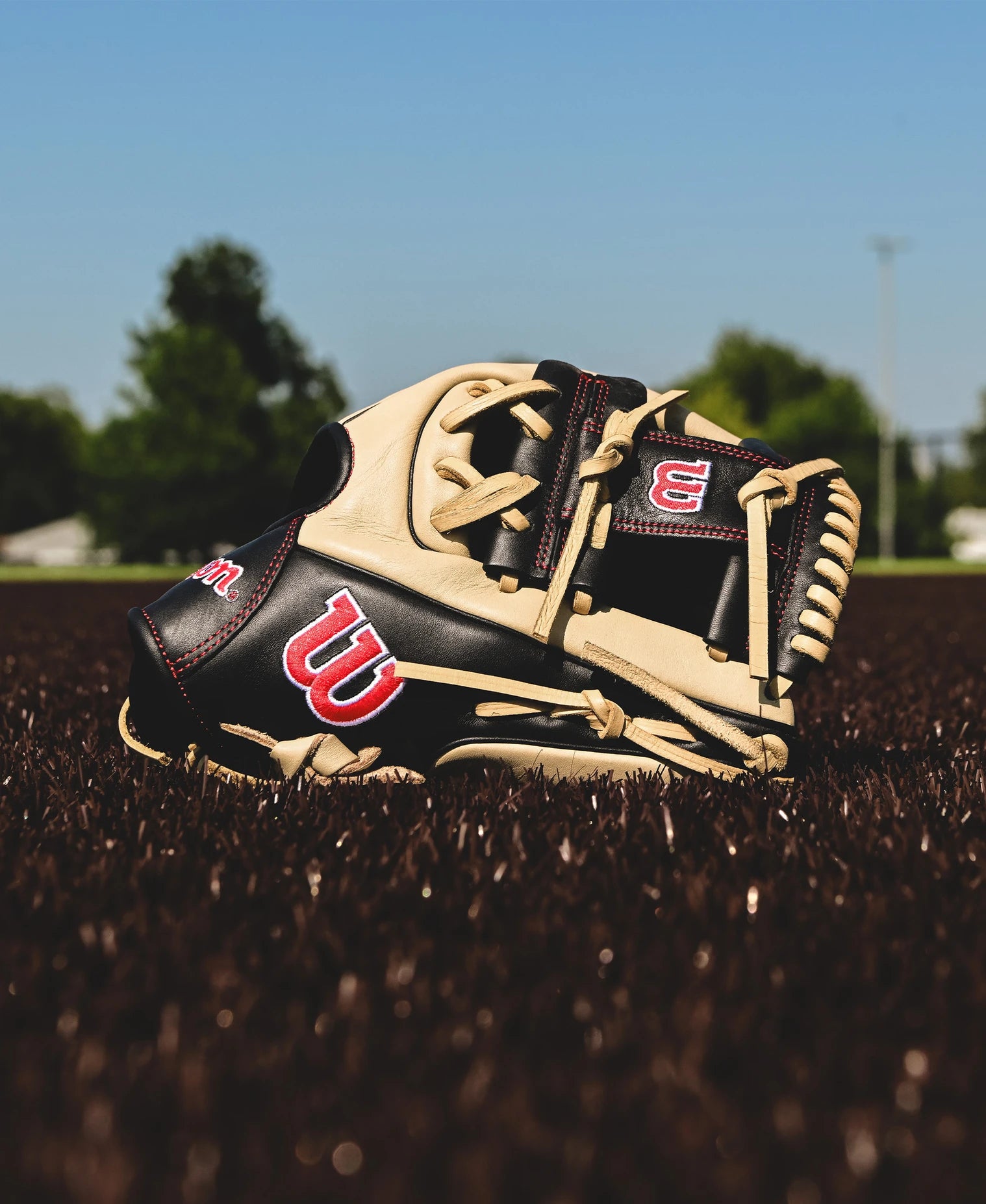 A Wilson A900 PF115 11.5" Baseball Glove (WBW102570115) with black and tan leather sits on brown artificial turf, framed by trees and a clear blue sky.