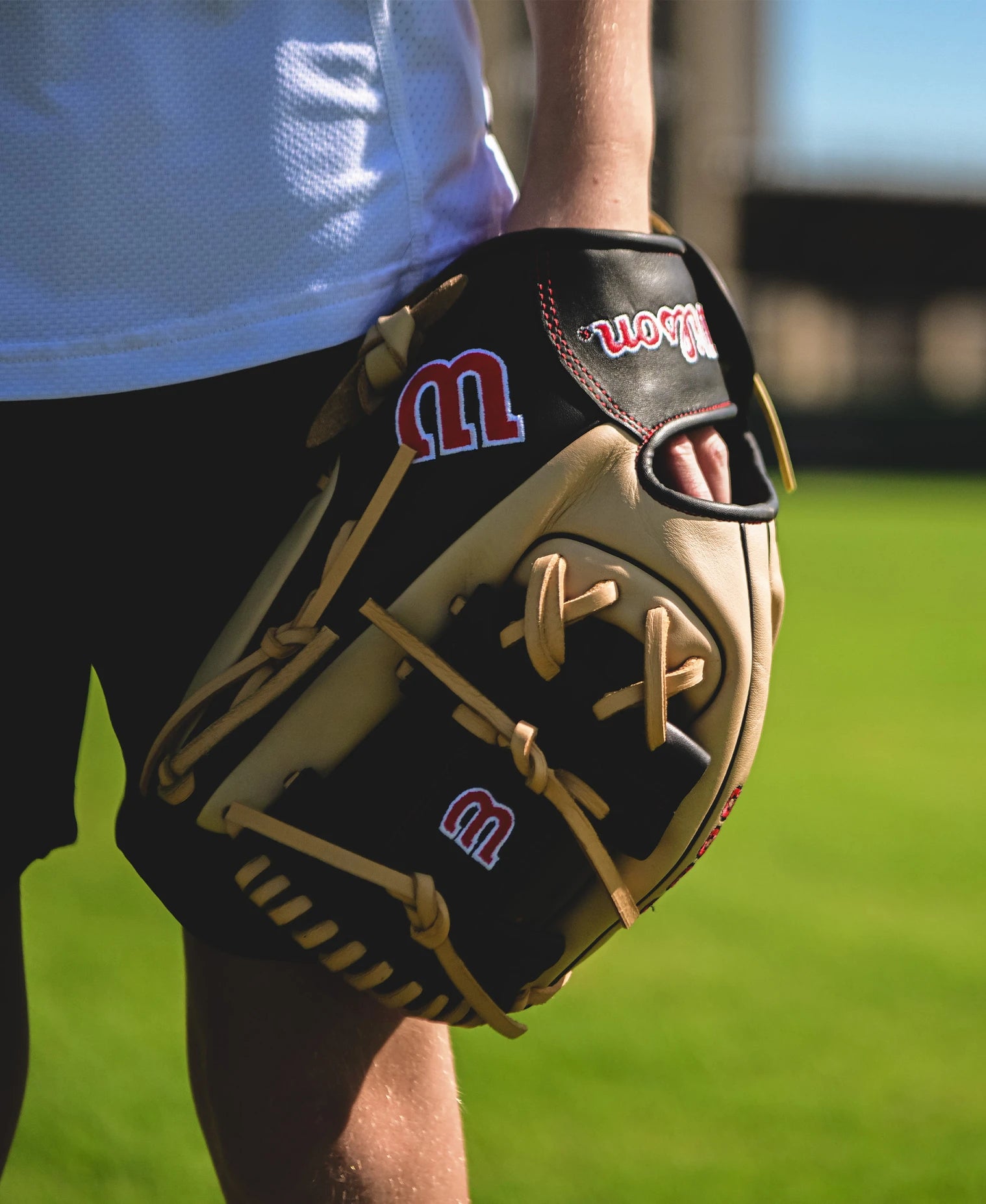 A person holds a tan and black Wilson A900 PF115 11.5" Baseball Glove (WBW102570115) on a sunny field with green grass, wearing a white shirt and black shorts.