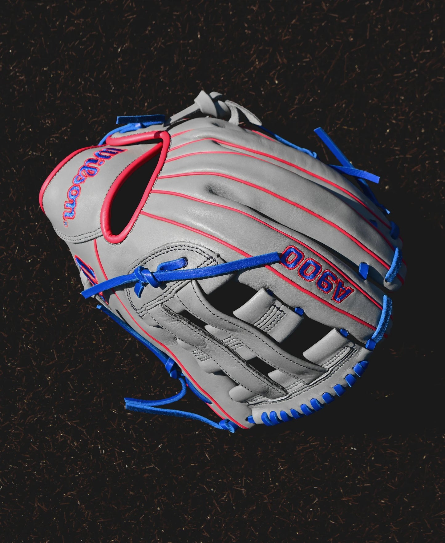 A Wilson A900 PF12 12" Baseball Glove (WBW10257112) in gray with red stitching and blue laces rests on a dark surface, ideal as a youth baseball glove.