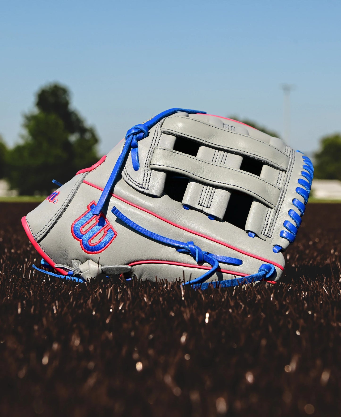 A Wilson A900 PF12 12" baseball glove (WBW10257112) in gray with blue laces and pink accents lies on brown artificial turf, set against trees and a blue sky.