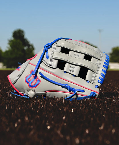 A Wilson A900 PF12 12" baseball glove (WBW10257112) in gray with blue laces and pink accents lies on brown artificial turf, set against trees and a blue sky.