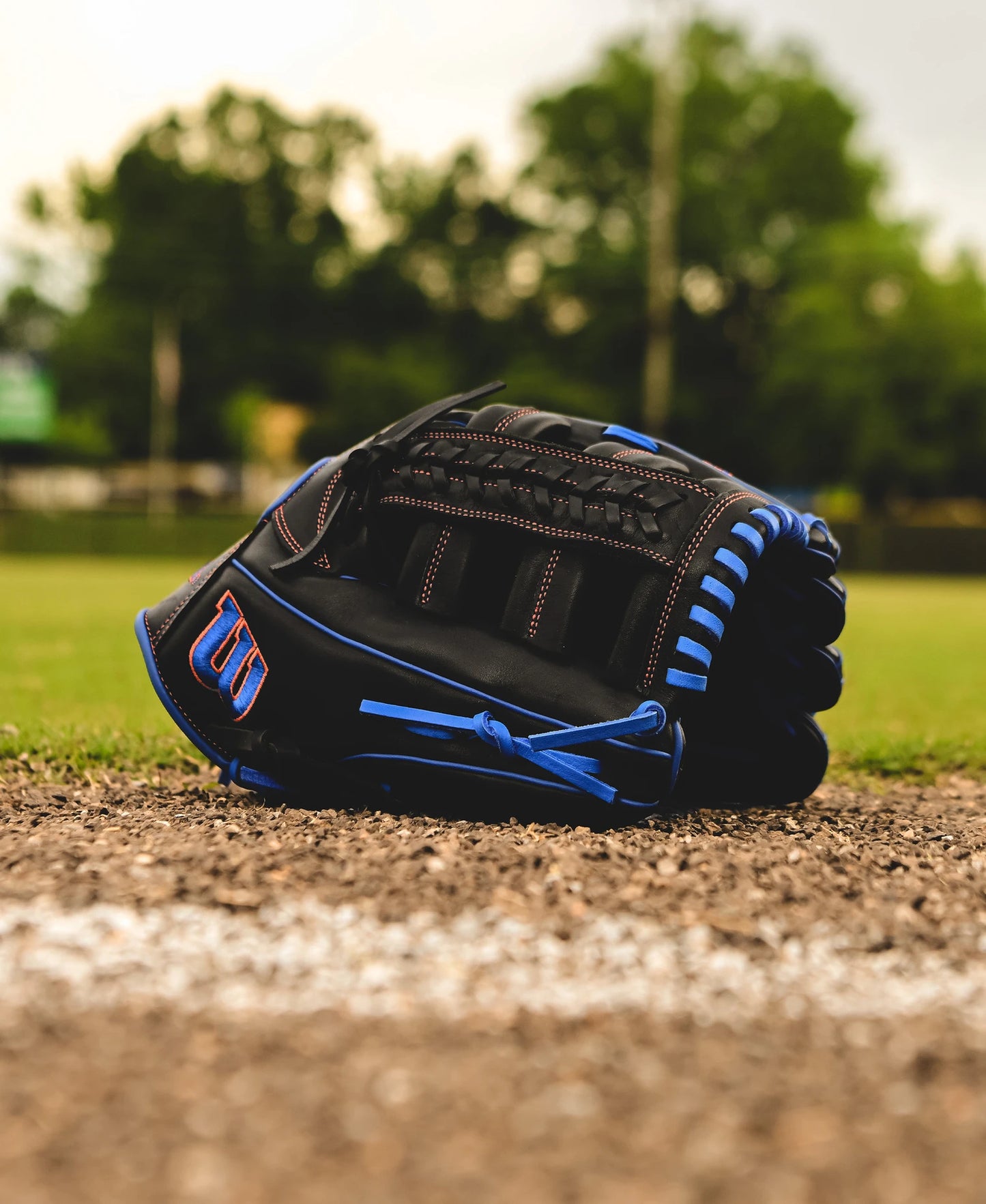 A Wilson A1000 PF1892 12.25" Baseball Glove (WBW1025831225) rests on the ground near the edge of a baseball field, framed by green trees and a softly blurred background.