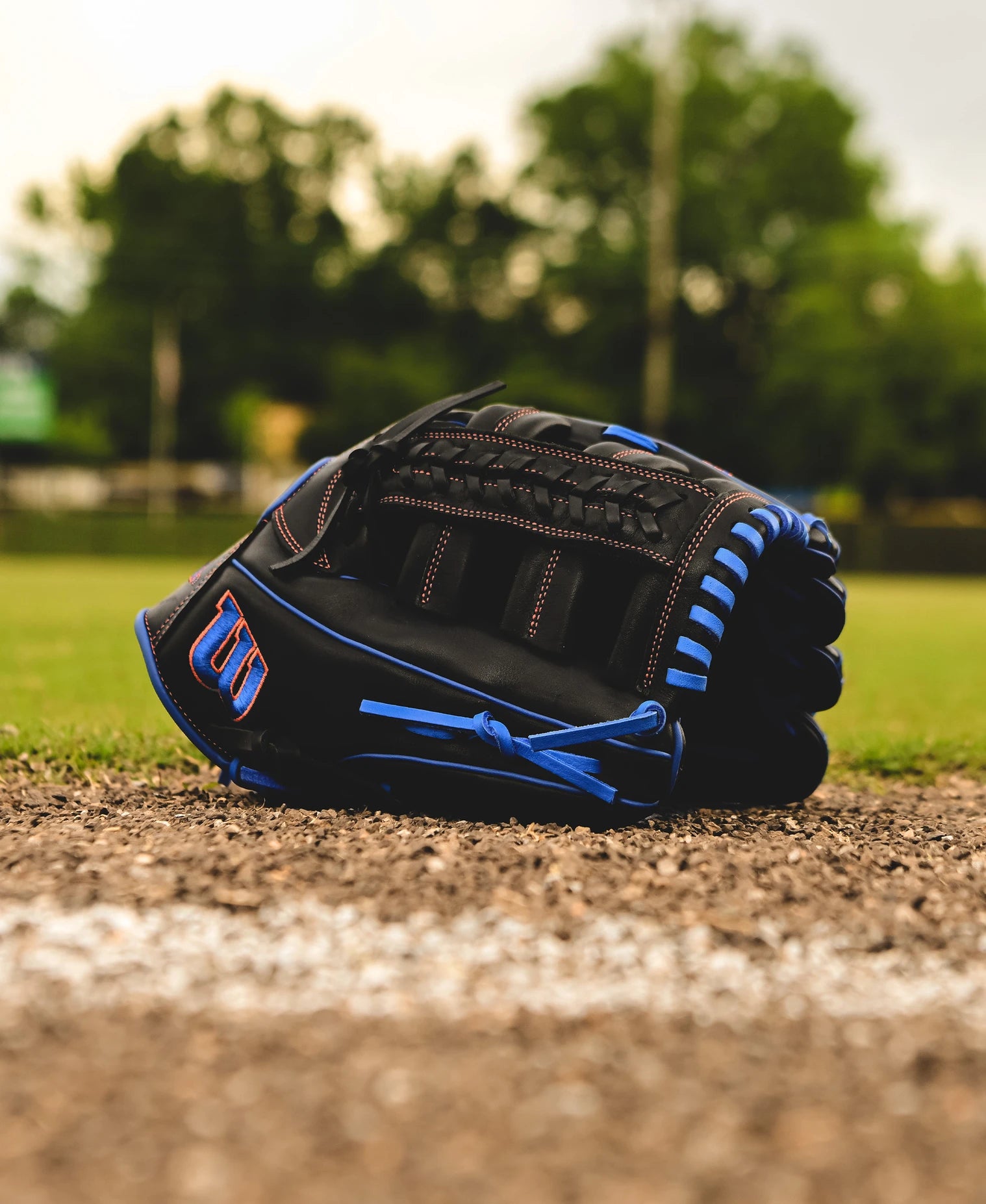 A Wilson A1000 PF1892 12.25" Baseball Glove (WBW1025831225) rests on the ground near the edge of a baseball field, framed by green trees and a softly blurred background.