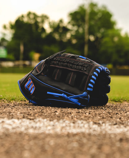 A Wilson A1000 PF1892 12.25" Baseball Glove (WBW1025831225) rests on the ground near the edge of a baseball field, framed by green trees and a softly blurred background.
