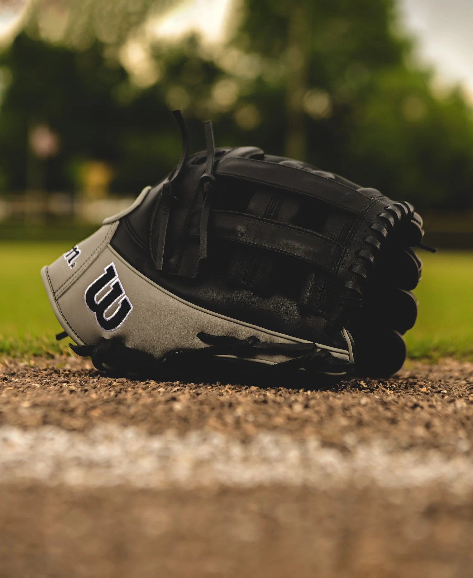 A Wilson A1000 1750 12.5" Baseball Glove (WBW102585125) in black and gray Full Grain leather rests on the dirt by a white chalk baseline, with an outdoor field, green trees, and a blurred background behind it.