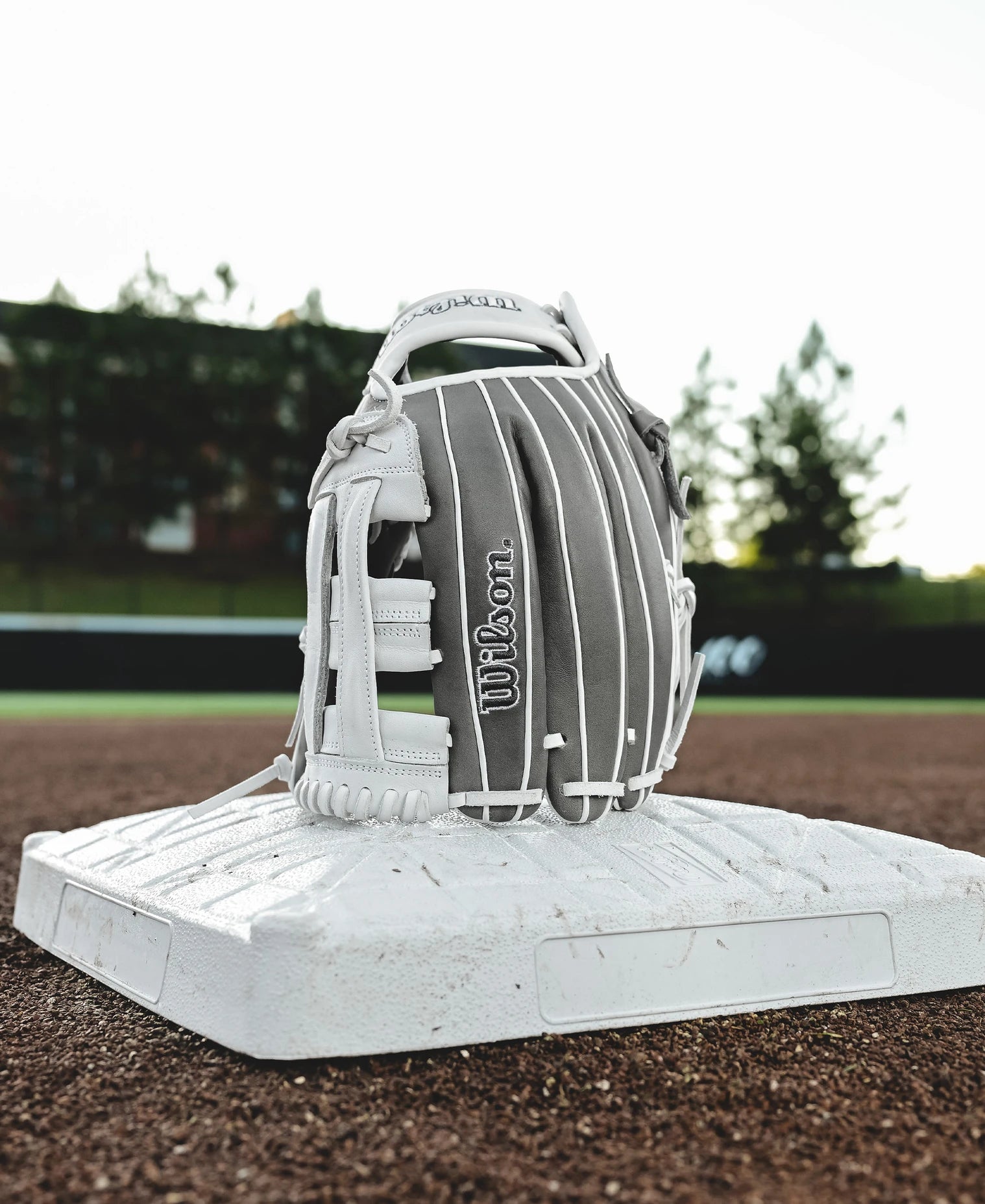 A Wilson A1000 IF12 12" Fastpitch Glove (gray/white), crafted from Full Grain leather, stands upright on a white base on a baseball field, with grass, dirt, and trees in the background.
