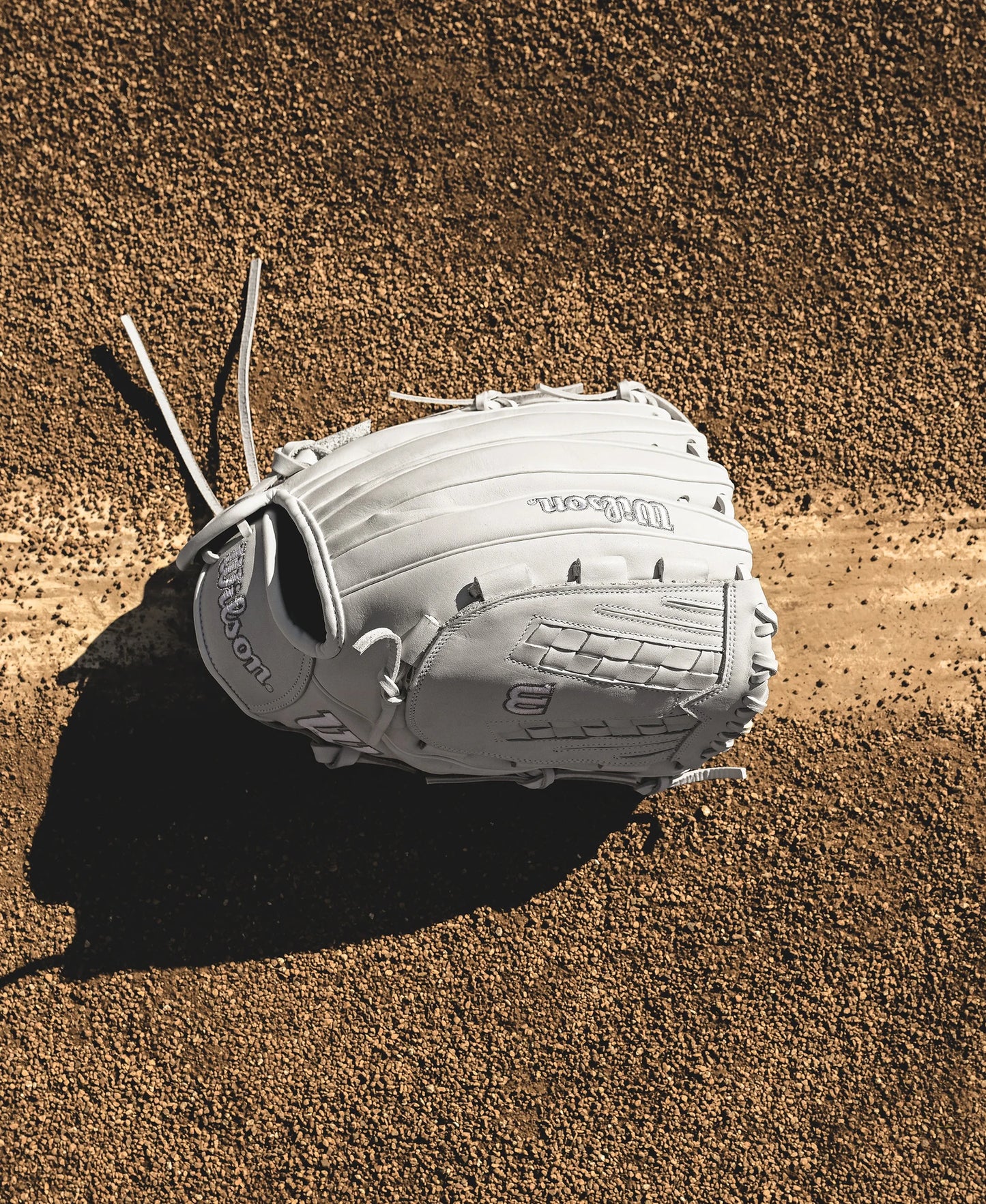A Wilson A1000 V125 12.5" Fastpitch Glove (WBW102594125) rests on dirt, partly covering a bright white foul line, casting a shadow to the left on the textured field.