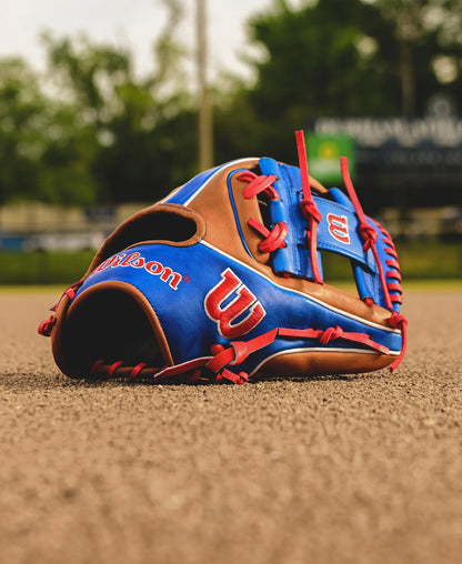 A Wilson A2K 1786 11.5" Mookie Betts GM Baseball Glove in blue, brown, and red Pro Stock Select leather rests on a baseball field’s dirt, with blurred trees and a scoreboard in the background.