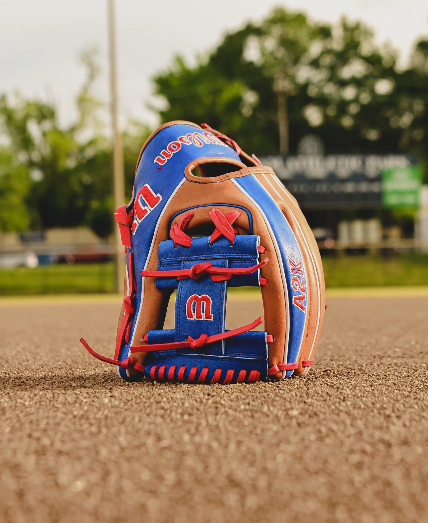The Wilson A2K 1786 11.5" Mookie Betts GM Baseball Glove (FALL 2024) features red, blue, and tan Pro Stock Select leather, standing upright on an infield with trees and a blurred scoreboard in the background.