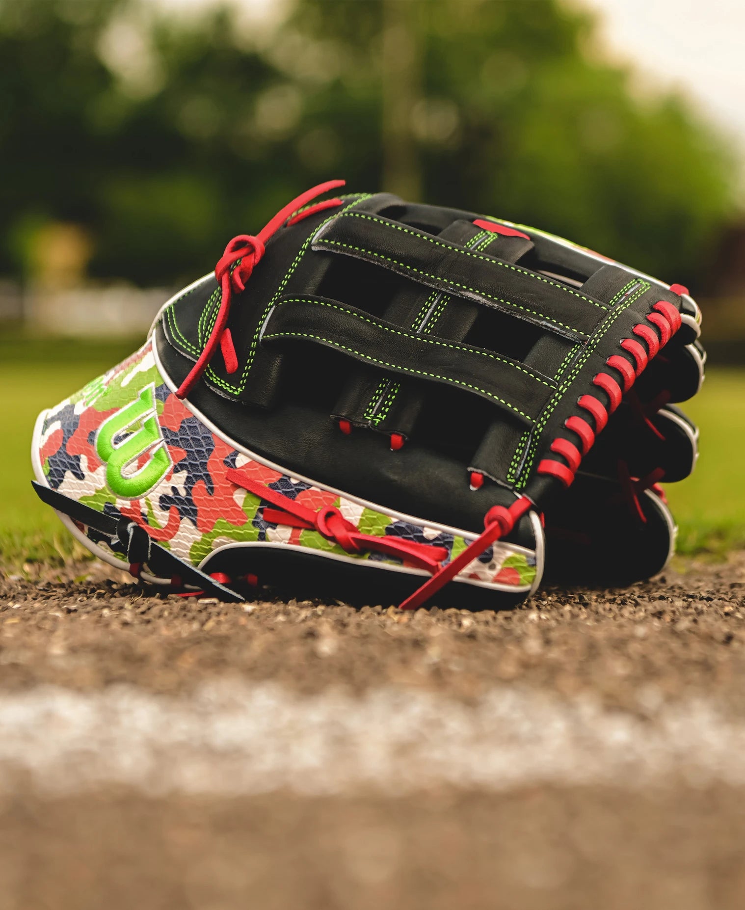 A Wilson A2K MH23 12.75" Michael Harris GM Baseball Glove, featuring red laces and camouflage patterns, rests near the edge of a dirt baseball field with green grass and blurred trees in the background.