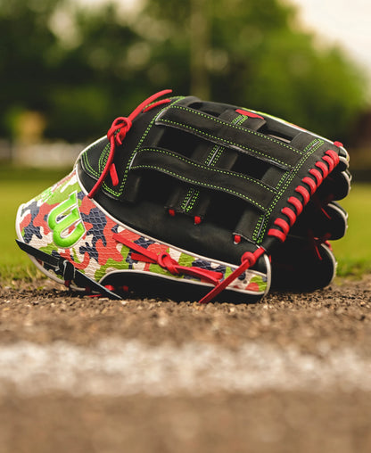 A Wilson A2K MH23 12.75" Michael Harris GM Baseball Glove, featuring red laces and camouflage patterns, rests near the edge of a dirt baseball field with green grass and blurred trees in the background.