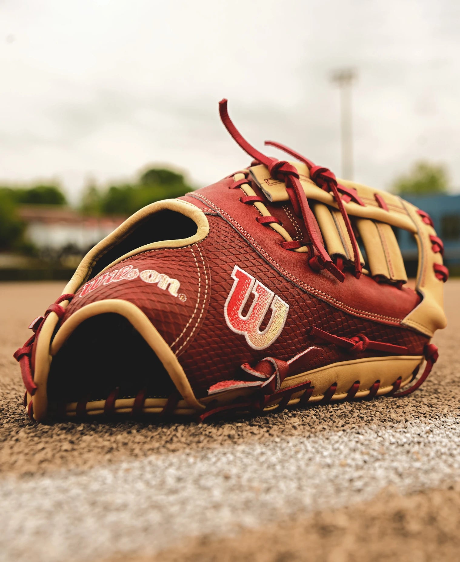 A Wilson A2K 1679 12.5" Baseball First Base Mitt (FALL 2024: WBW102684125) in red and tan leather rests on a dirt field by a chalk line, with blurred trees and sky in the background.