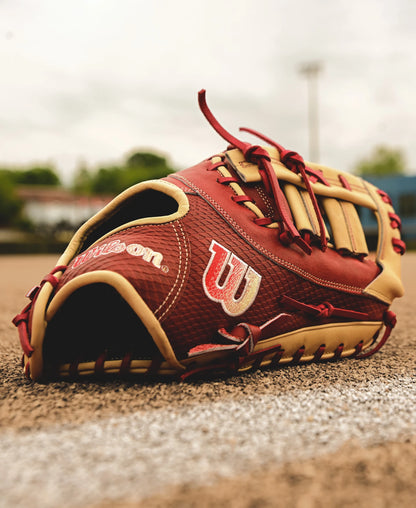 A Wilson A2K 1679 12.5" Baseball First Base Mitt (FALL 2024: WBW102684125) in red and tan leather rests on a dirt field by a chalk line, with blurred trees and sky in the background.